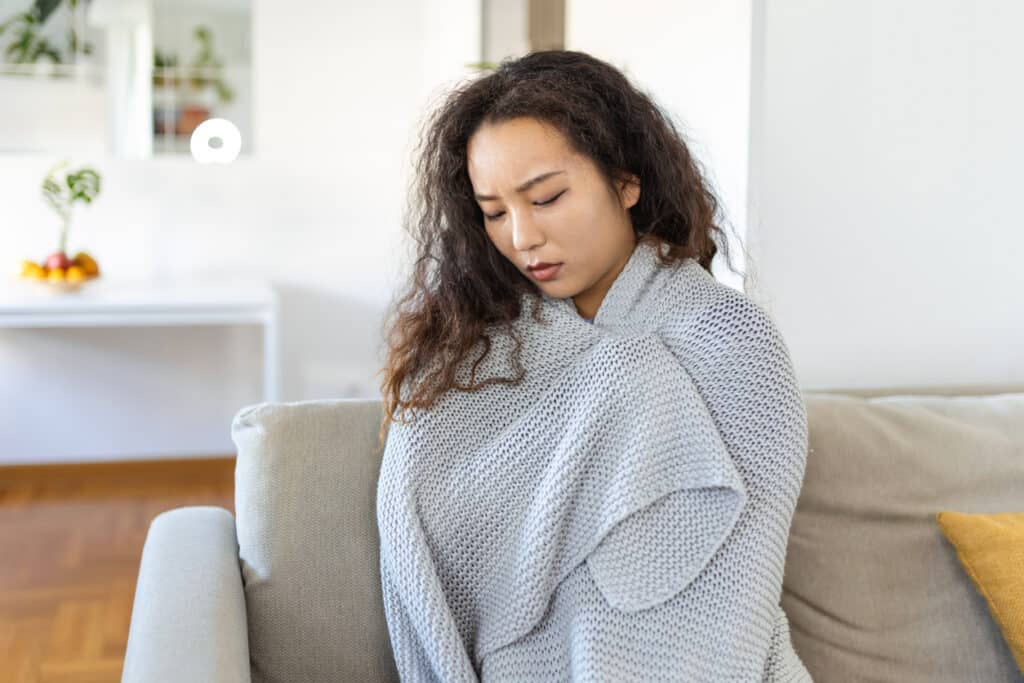 Sick young Asian woman feeling cold covered with blanket sit on bed, ill black girl shivering freezing warming at home wrapped with plaid, no central heating problem, fever temperature flu concept in spring time