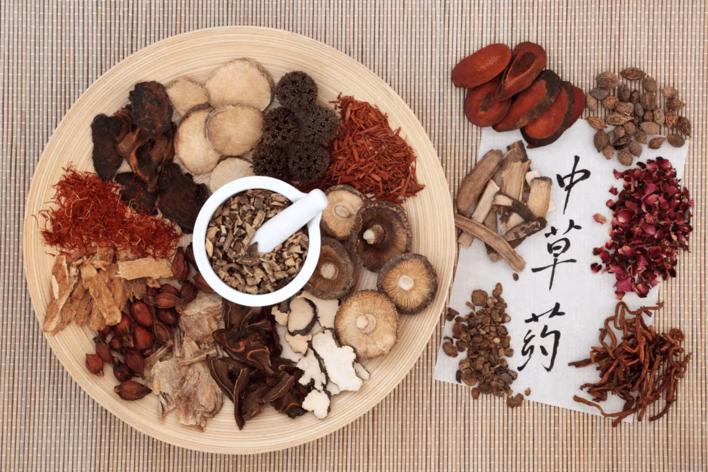 Traditional Chinese medicine herbs, dried roots, mushrooms, flowers, and a mortar arranged on a bamboo mat