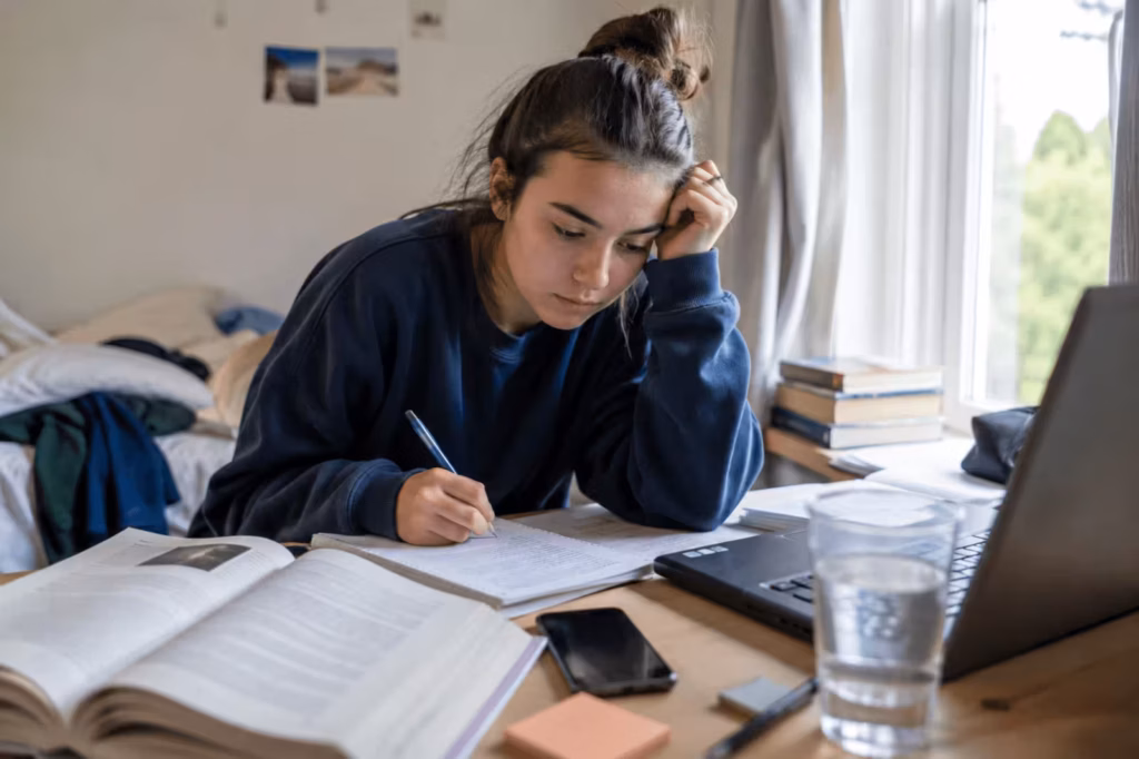 Tired student studying at a desk with books, laptop, and phone